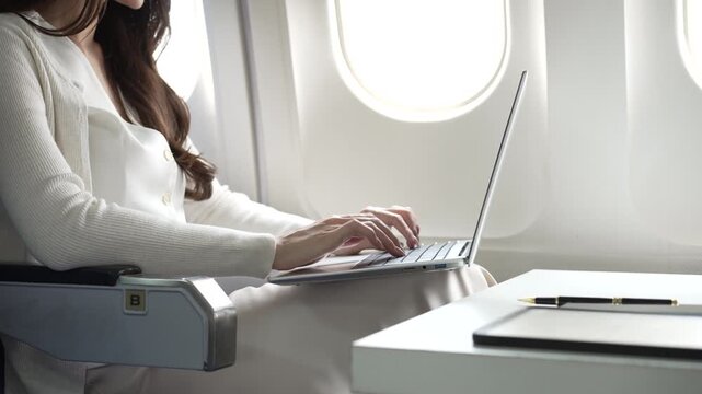 Business traveler working on laptop in airplane cabin by window, with notebook and pen on tray table, bright daylight, modern travel and remote work concept.