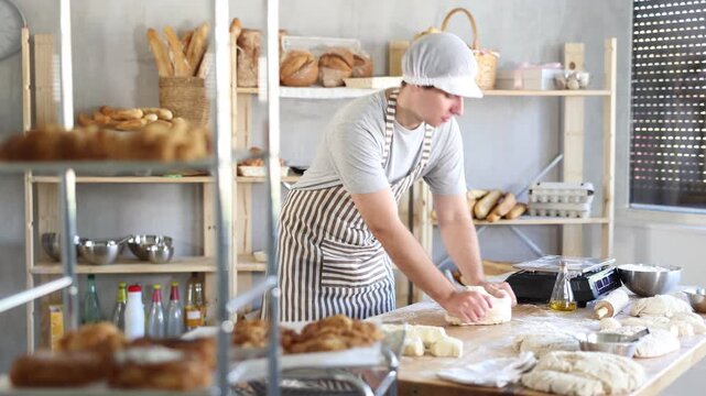 Young male baker stands at his work bench, kneading and shaping dough to make bread, croissants and baguettes