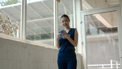 Fototapeta premium Smiling young businesswoman in blue suit using smartphone in bright modern office corridor with large windows, natural light, casual workplace communication and technology concept.