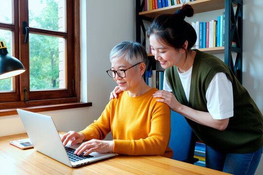Asian senior woman using laptop while young adult daughter assisting at home, showing digital learning and family support. Useful for tech training, caregiving, online services marketing