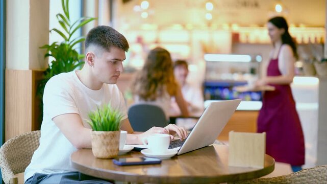 Young man is sitting at a table in a cafe and working at a laptop. Guy designer works remotely in a coffee shop. High quality 4k footage