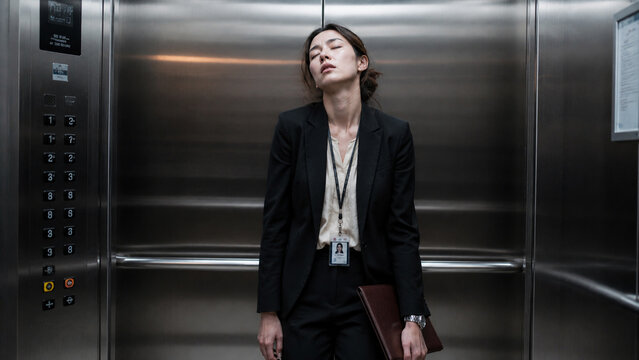 An exhausted businesswoman leans against the elevator wall, overwhelmed by a demanding workday, reflecting the silent struggle and fatigue of corporate life