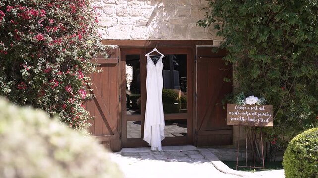 A wedding dress hangs from a wooden door at a venue. Beside it is a sign with a quote. Flower bushes frame the setting. The location looks nice and inviting for guests.