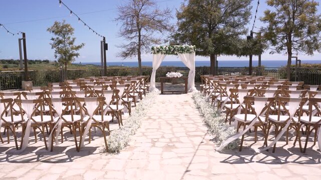 A wedding setup is ready with rows of empty chairs facing an altar. The ocean can be seen in the background. The sun shines brightly, and trees line the area.