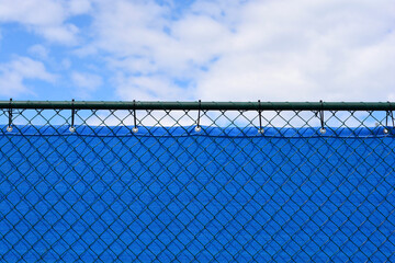 Fototapeta premium An eye level shot shows the top edge of a green wire mesh fence lined with a bright blue privacy screen against a partly cloudy sky.