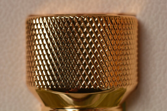 A macro close up view of a shiny golden metal handle featuring a diamond knurled pattern texture against a soft beige background