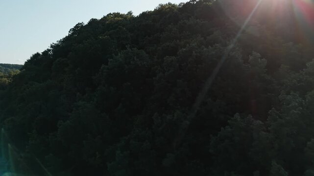 Sunlight over dense forested hilltop patch, silhouette backlit medium aerial ascend