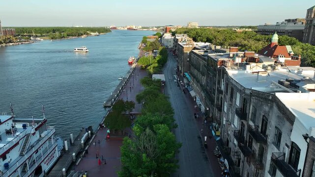 Drone footage of Savannah, Georgia, capturing the riverfront promenade, historic district, and a riverboat docked beside the bustling cobblestone streets.