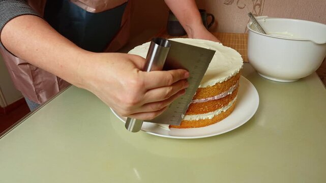 A close-up shot shows a baker meticulously smoothing the white frosting on a tall, layered cake using a metal bench scraper for a professional finish