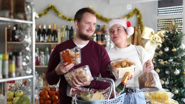  Man and a woman in a Santa hat holding delicious food in a New Year's supermarket. Happy couple buying mandarins and sweets for Christmas