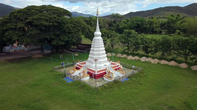 A smooth aerial drone orbit around a beautiful white pagoda with golden Naga statues at its base, surrounded by lush green fields and mountains.