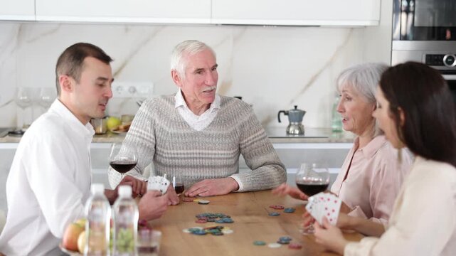Joyful senior parents and young couple sitting around table playing with cards and drinking wine in goblets at home