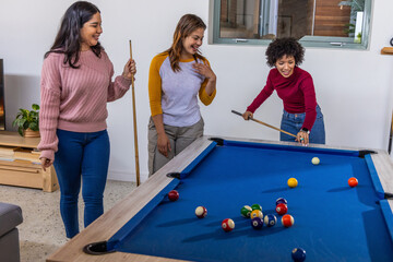 Fototapeta premium Diverse female friends leaning, watching, lining up shot on blue-felt pool table with cues at home