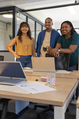 Fototapeta premium Diverse coworkers smiling and standing in modern office at wooden table with laptops, pie chart