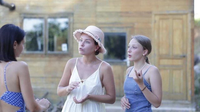 On bright summer day, three young European women neighbors quarrel and swear rural yard. Woman stopped near house and screams and insults stubborn girl, reproaches and accuses