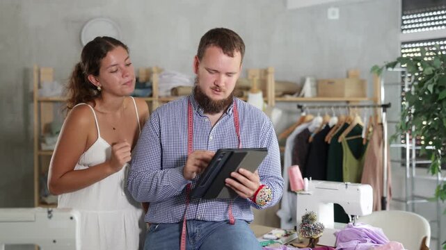 Careful young male dressmaker discussing dress design on tablet with girl in fashion studio . High quality 4k footage