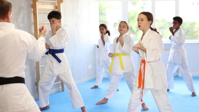 Focused tween girl, karateka beginner in kimono, standing in attacking stance, practicing punches or hand striking techniques as part of kata with group of teenagers