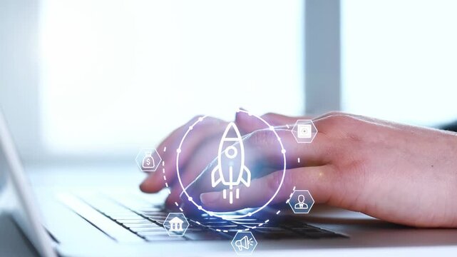 Person types on a laptop while graphic elements of a rocket and icons are visible on the screen in a modern workspace setting.