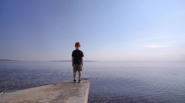 A young boy in a vibrant red cap stands on a concrete pier, looking out at the boundless water. His quiet posture reflects a moment of deep contemplation under a vast, clear blue sky.