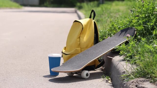 Yellow backpack, skateboard and coffee near the curb on a background of green grass. Concept of active lifestyle, youth recreation and summer vacation. Morning in the city, skateboard and coffee
