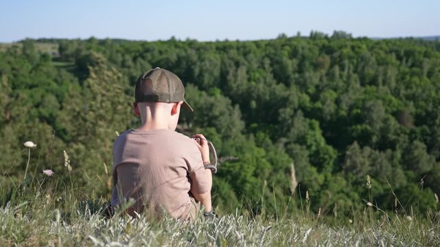 Lonely young boy wearing a cap sitting on the edge of a grassy hill, peacefully looking out over a vast green forest on a sunny day, representing a moment of quiet contemplation