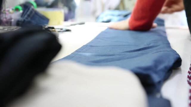 의류 봉제 공장 작업실 옷 수선 바지 재단 원단 쓸어내리기 자재: 빨간색 상의를 입은 숙련공이 작업대 위에 어두운 남색 바지 원단을 길게 펼쳐놓고 손으로 표면을 평평하게 매만지며 정돈하는 모습을 포착한 클로즈업 영상입니다. 일터의 현장감을 담았으며, 패션 디자인 의류 제조, 섬유 가공 공정, 옷 수선 리폼 및 소상공인 생산 콘셉트의 실감 나는 인서트 숏을 