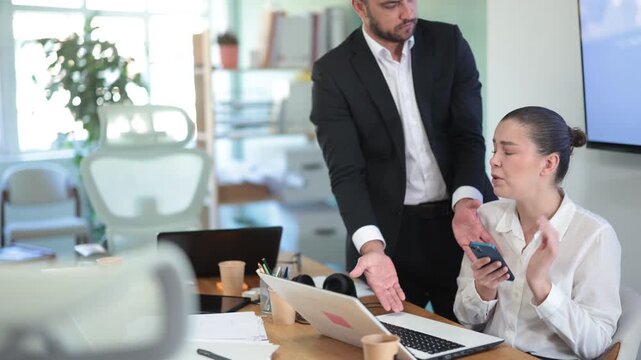 Young man, the boss, scolds at a woman who sits at the table and scrolls through social networks on mobile phone instead of working. Man expresses his dissatisfaction to an employee in the office