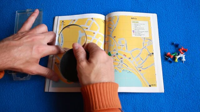Lupa y libro callejero buscando direcci&oacute;n mapa con lente de aumento y mano con dedo se&ntilde;alando ruta, punto de encuentro y destino en hoja p&aacute;gina de papel