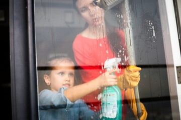 Fototapeta premium Woman and child cleaning a window with spray and squeegee indoors.