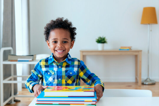Portrait of Black boy smiling at camera while holding stack of books at table, suggesting early learning and reading success. Useful for education, literacy, child development campaigns