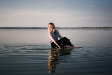 Fototapeta premium Portrait of beautiful young woman in dress sits in the water doing circles on water. Copy space. Fashion concept. Energy of water, earth and sun. Witch ritual. State of pleasure and bliss. Sexy pose.