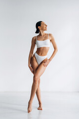 Fototapeta premium Slim female model in white lingerie poses gracefully against a plain white background, showcasing a toned physique and elegant posture with soft lighting