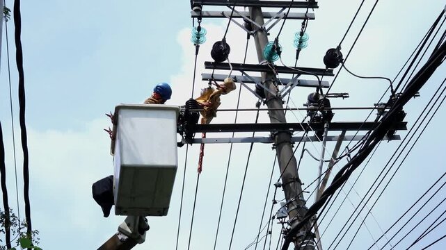Electrician works on a tall pole. He climbs high above tangled wires. The worker uses a bucket truck for access. He wears protective gear and safety helmet. The sky is clear and blue behind him
