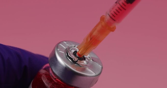 Syringe needle penetrating the rubber stopper of a medical vial held by a hand in a detailed view on a pink gradient background.
