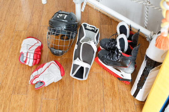 Hockey gear is arranged on a wooden floor in a training space. Items include a helmet, skates, gloves, and protective padding, showing readiness for practice or a game.