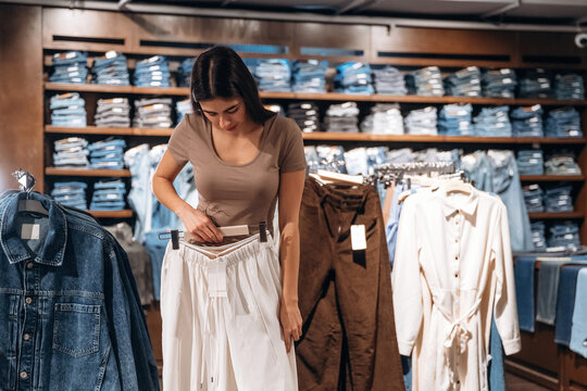 Female shopper examining white pants while standing in a clothing store with shelves of folded denim and garments in the background