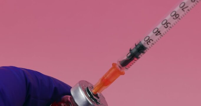 A hand holding a pharmaceutical glass vial with a syringe needle inserting into the rubber top on a pink gradient background.