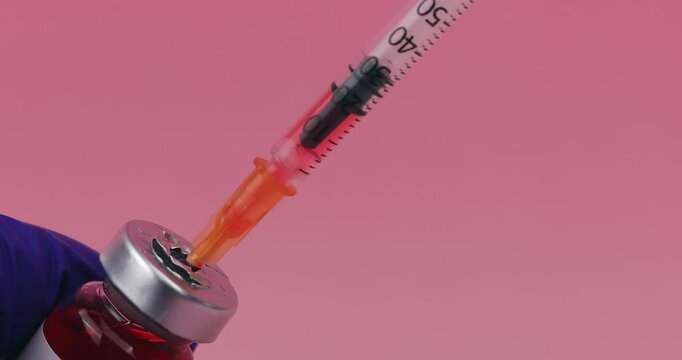 Hand holding a medical glass vial while a syringe needle inserts into the rubber stopper over a beautiful pink gradient background.