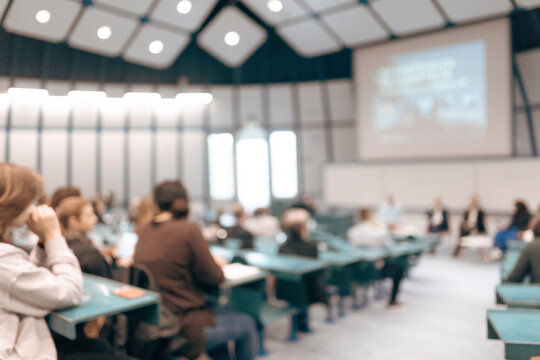 A large group of diverse people attending a professional presentation or educational workshop in a modern lecture hall. Defocus image of audience from behind.