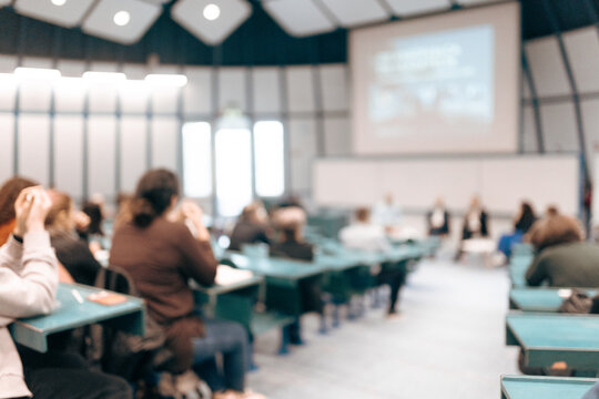 A large group of diverse people attending a professional presentation or educational workshop in a modern lecture hall. Defocus image of audience from behind.
