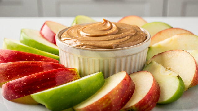 Healthy snack platter featuring a ceramic bowl of smooth creamy peanut butter surrounded by fresh red and green apple slices.