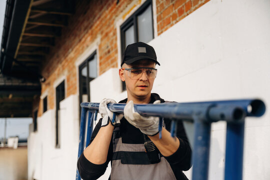 Male construction worker in gray overalls and safety glasses carrying a blue scaffolding pipe outside a building with brick walls and windows