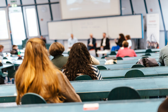 A large group of diverse people attending a professional presentation or educational workshop in a modern lecture hall. Focus on the audience from behind.