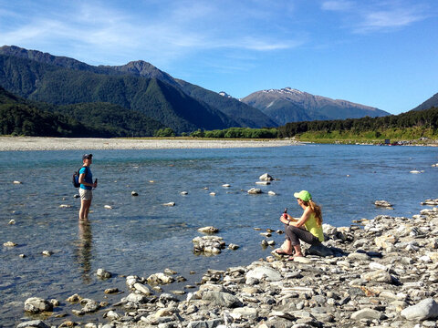Two caucasian women chatting and drinking beer in river with mountains in background.