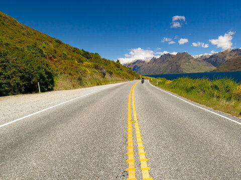 Looking along centre double line of a road beside a lake with a motorbike rider