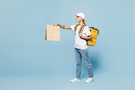 Full body employee delivery woman wear white T-shirt uniform thermal food backpack work as courier hold brown blank craft paper takeaway packet bag isolated on plain blue background. Service concept.