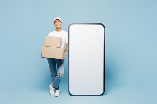 Full body delivery employee woman wears white cap t-shirt uniform workwear work as dealer courier big huge blank screen mobile cell phone hold stack cardboard boxes isolated on plain blue background.