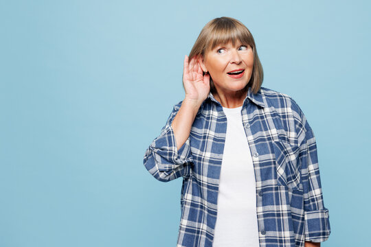 Elderly shocked curious nosy blonde woman 50s years old wear checkered shirt casual clothes try to hear you overhear listen intently isolated on plain pastel light blue background. Lifestyle concept.