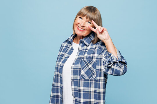 Elderly smiling happy positive blonde woman 50s years old wear checkered shirt casual clothes showing victory sign look camera isolated on plain pastel light blue background studio. Lifestyle concept.