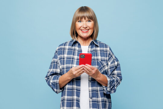 Elderly beautiful blonde woman 50s years old wear checkered shirt casual clothes hold in hand use mobile cell phone isolated on plain pastel light blue background studio portrait. Lifestyle concept.